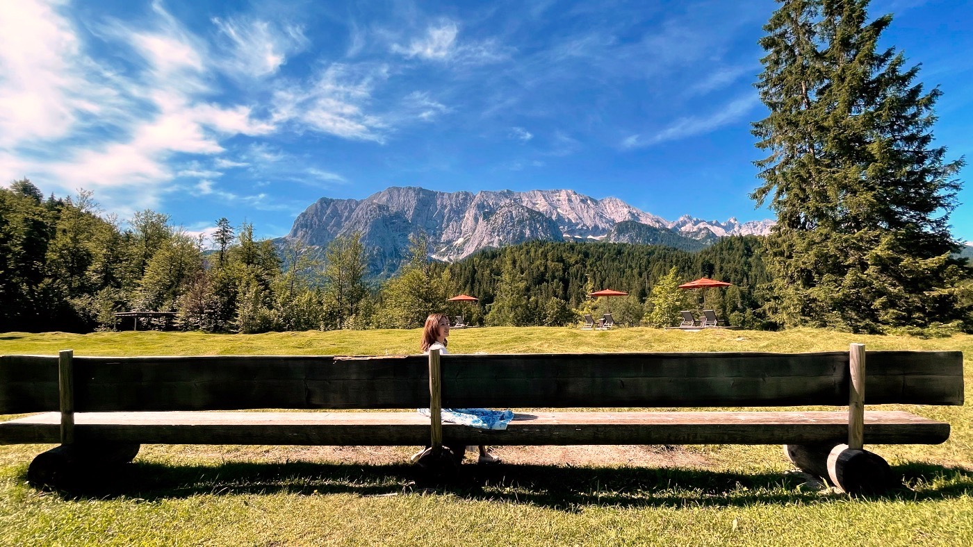 A woman on a long wooden bench in a meadow, the Wetterstein range and red parasols behind her