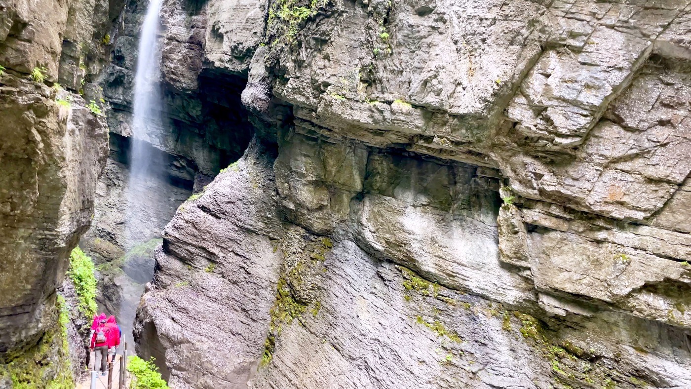 Hikers in red jackets on a narrow path beneath a waterfall in the Partnach Gorge
