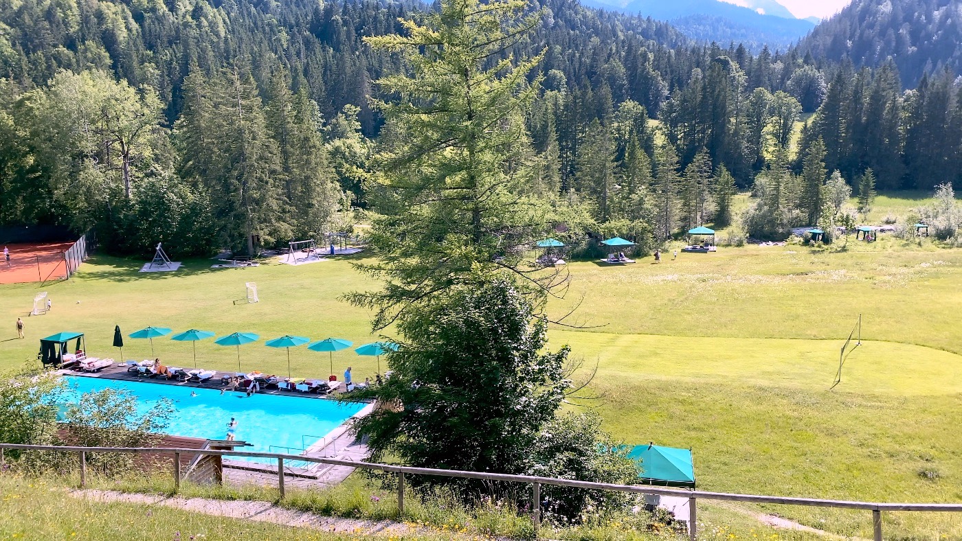 An outdoor pool with green parasols beside a wide meadow, ringed by forested alpine slopes