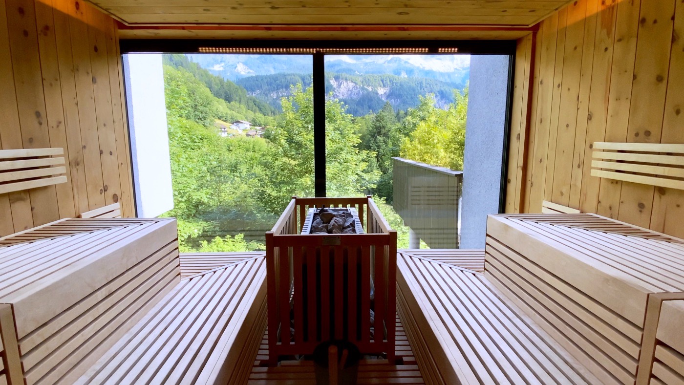 A pine sauna with a panoramic window framing the green alpine slopes