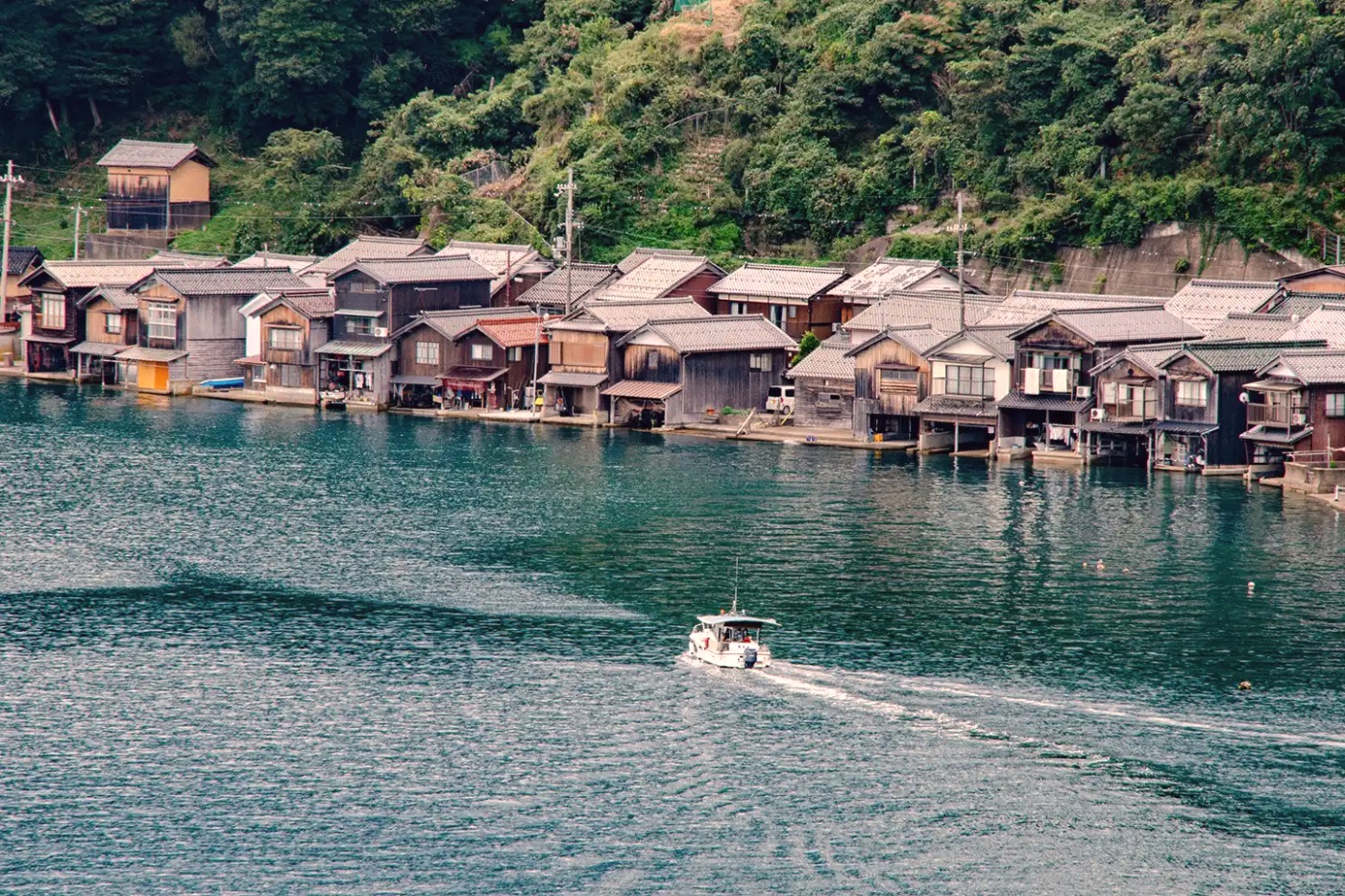 A row of Ine funaya boathouses lining the bay, a small boat cutting across the still water
