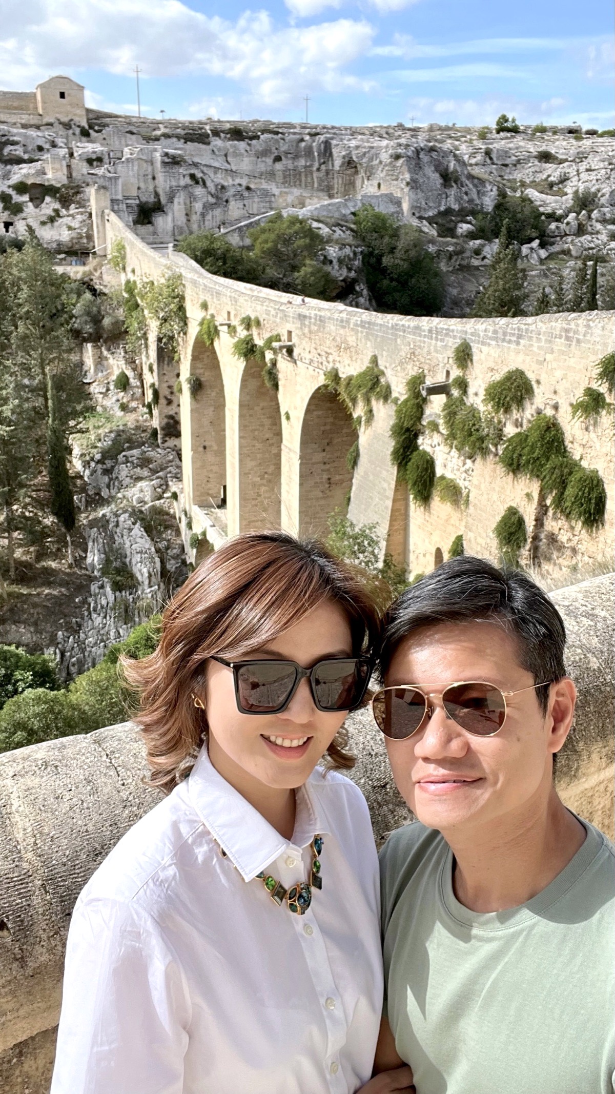 A couple in sunglasses posing in front of the arched Ponte Acquedotto over the Gravina gorge